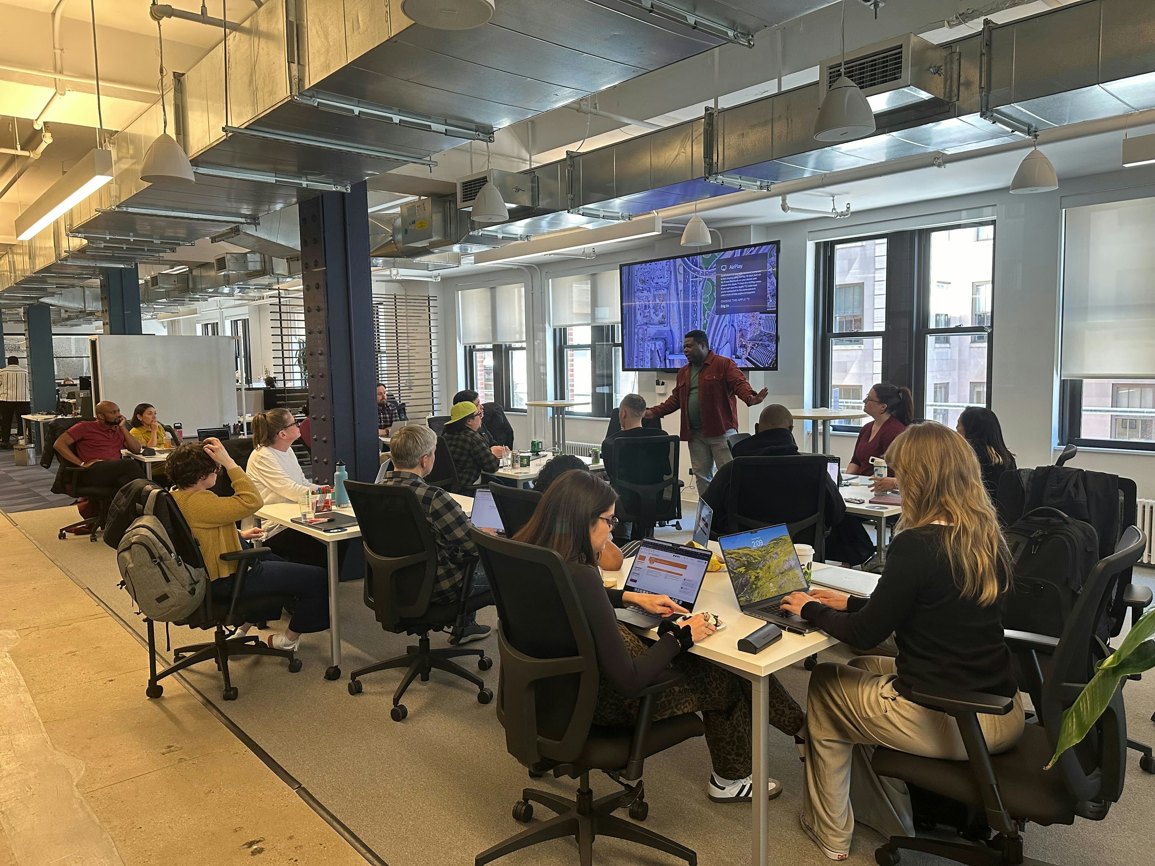 14 teammates sit in a conference space listening to one person standing in front of a large digital screen.